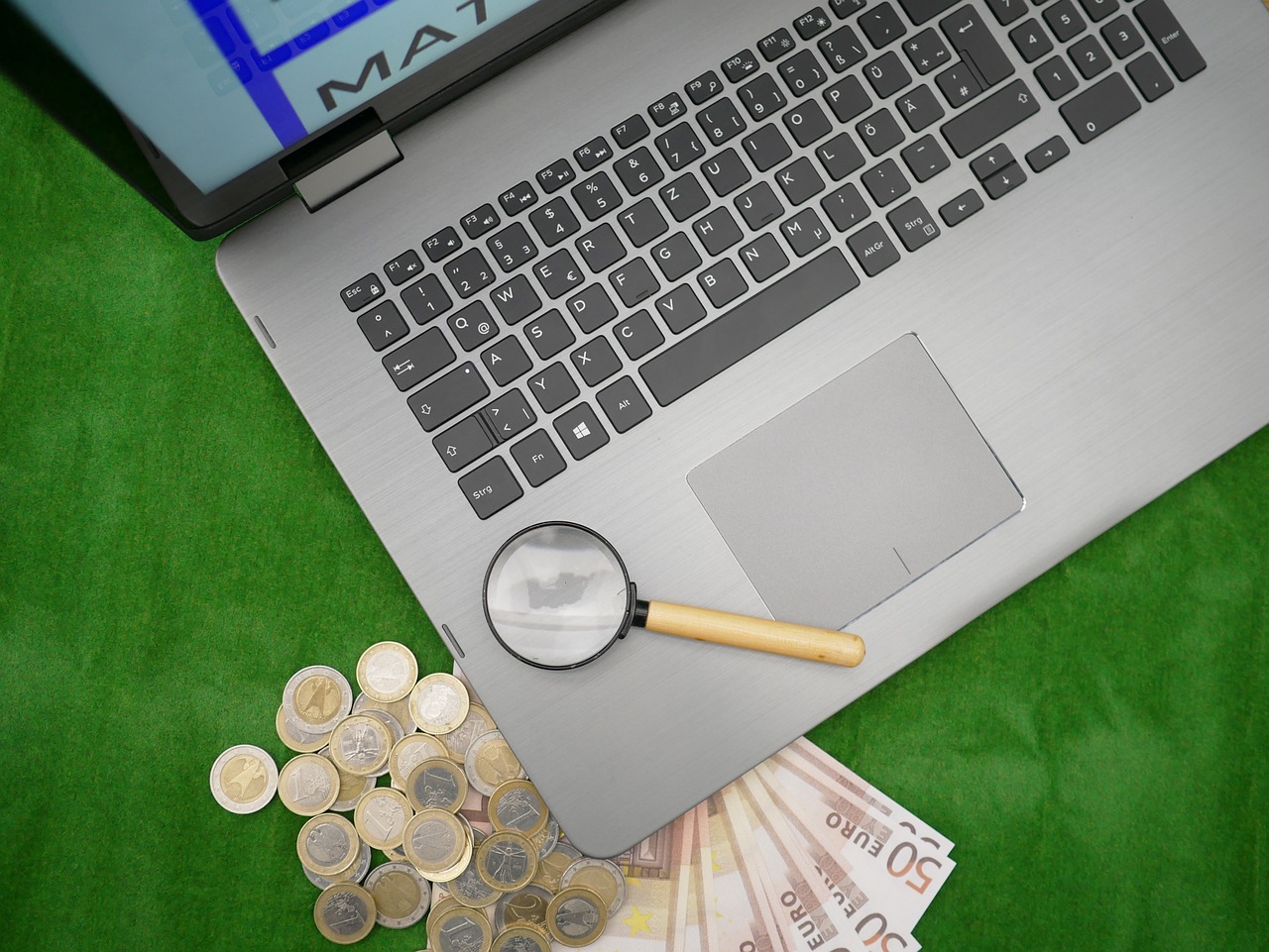 un laptop con una lente d'ingrandimento sulla tastiera, poggiato su un piano verde con alcune banconote e monete