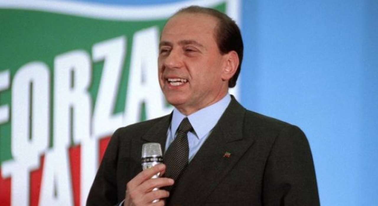 primo piano di Silvio Berlusconi con il microfono in mano e la bandiera di Forza Italia alle spalle
