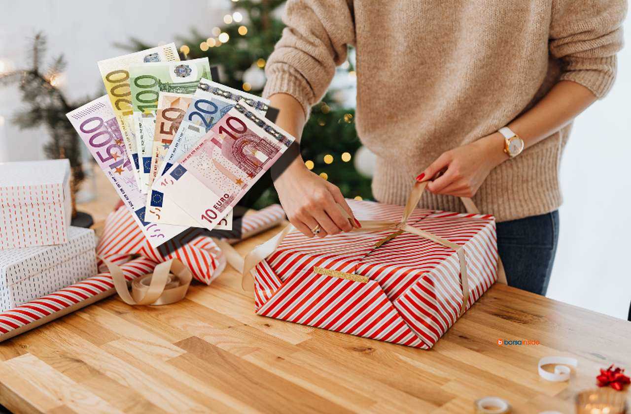 una donna che impacchetta un regalo con un albero di Natale sullo sfondo e alcune banconote di euro disposte a ventaglio in sovrimpressione