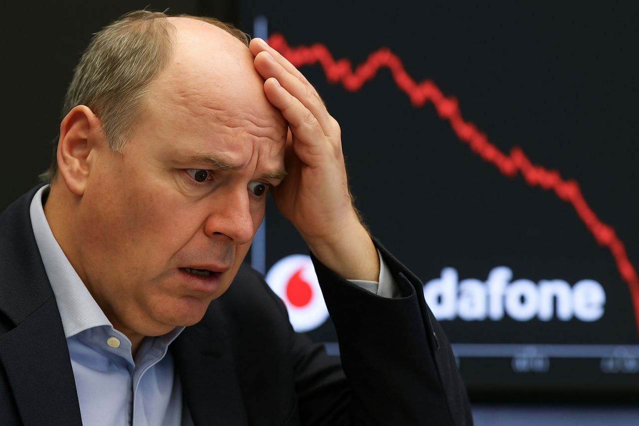 Primissimo piano di un uomo con una mano sulla fronte. Sullo sfondo un grafico finanziario in discesa con logo e scritta Vodafone