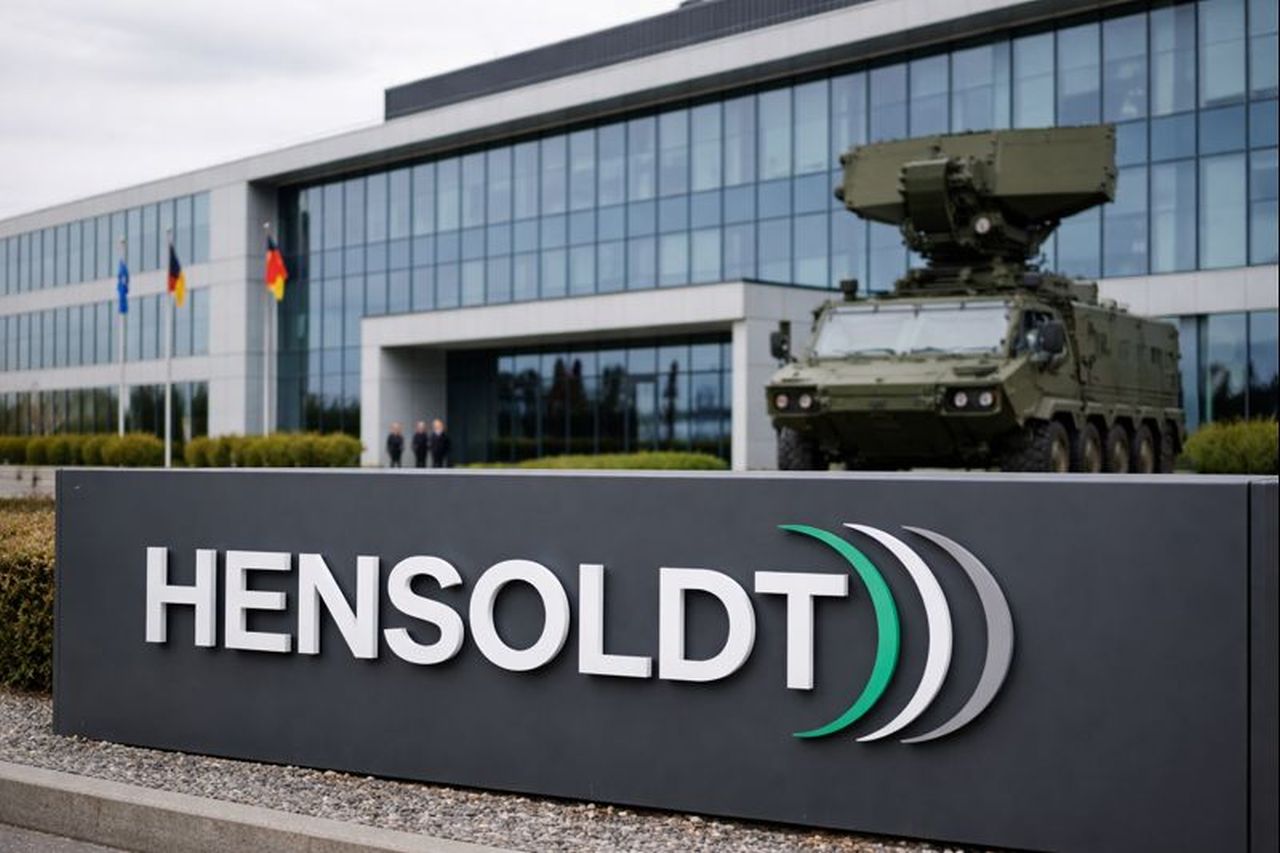 Logo Hensoldt in primo piano e un edificio con un mezzo militare parcheggiato davanti sullo sfondo