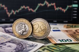 Moneta da una sterlina e moneta da due euro appoggiate su banconote euro e sterline, con un grafico finanziario sfocato sullo sfondo